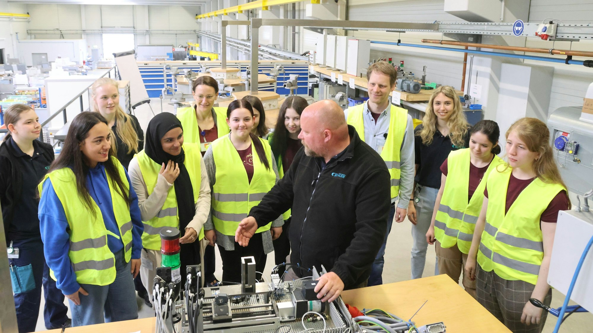 Eine Schülerinnengruppe steht in einer Firma und hört einem Leiter einer Führung durch die Produktion zu. Alle Mädchen tragen gelbe Warnwesten.