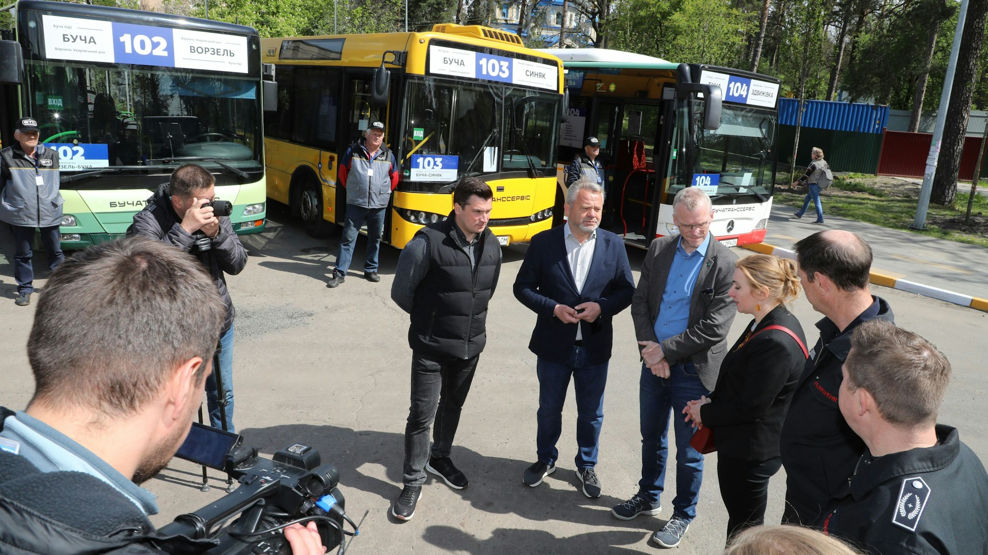 Menschen stehen im Halbkreis vor einer Reihe von Bussen. Ein Kameramann hält das Geschehen fest.