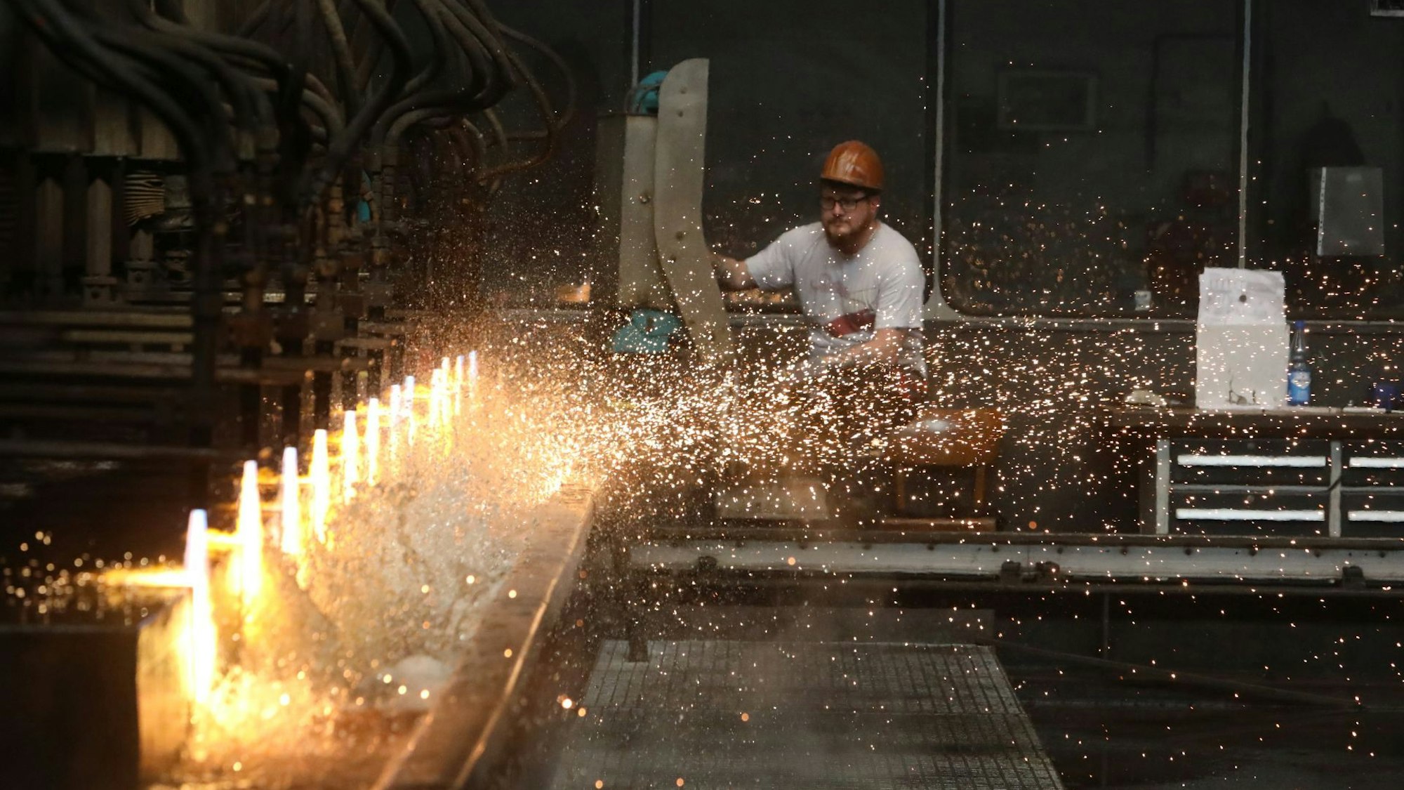Gelbe Flammen schneiden mit großer Hitze die Stahlplatten und lassen Wasser aufspritzen. Im Hintergrund bedient ein Mitarbeiter des Warmwalzwerks die Blockteilanlage.
