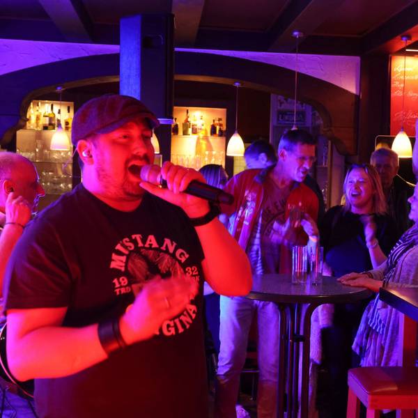 Ein Sänger einer Band singt in einer Kneipe. Im Hintergrund ist Publikum zu sehen.
