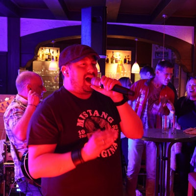 Ein Sänger einer Band singt in einer Kneipe. Im Hintergrund ist Publikum zu sehen.