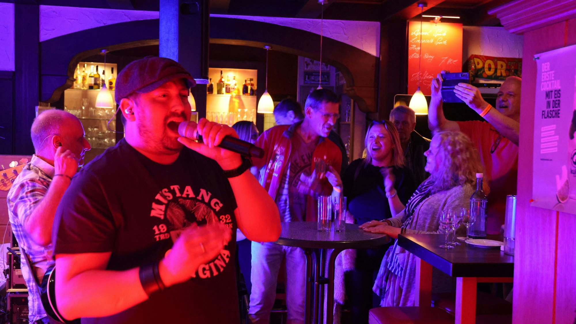 Ein Sänger einer Band singt in einer Kneipe. Im Hintergrund ist Publikum zu sehen.