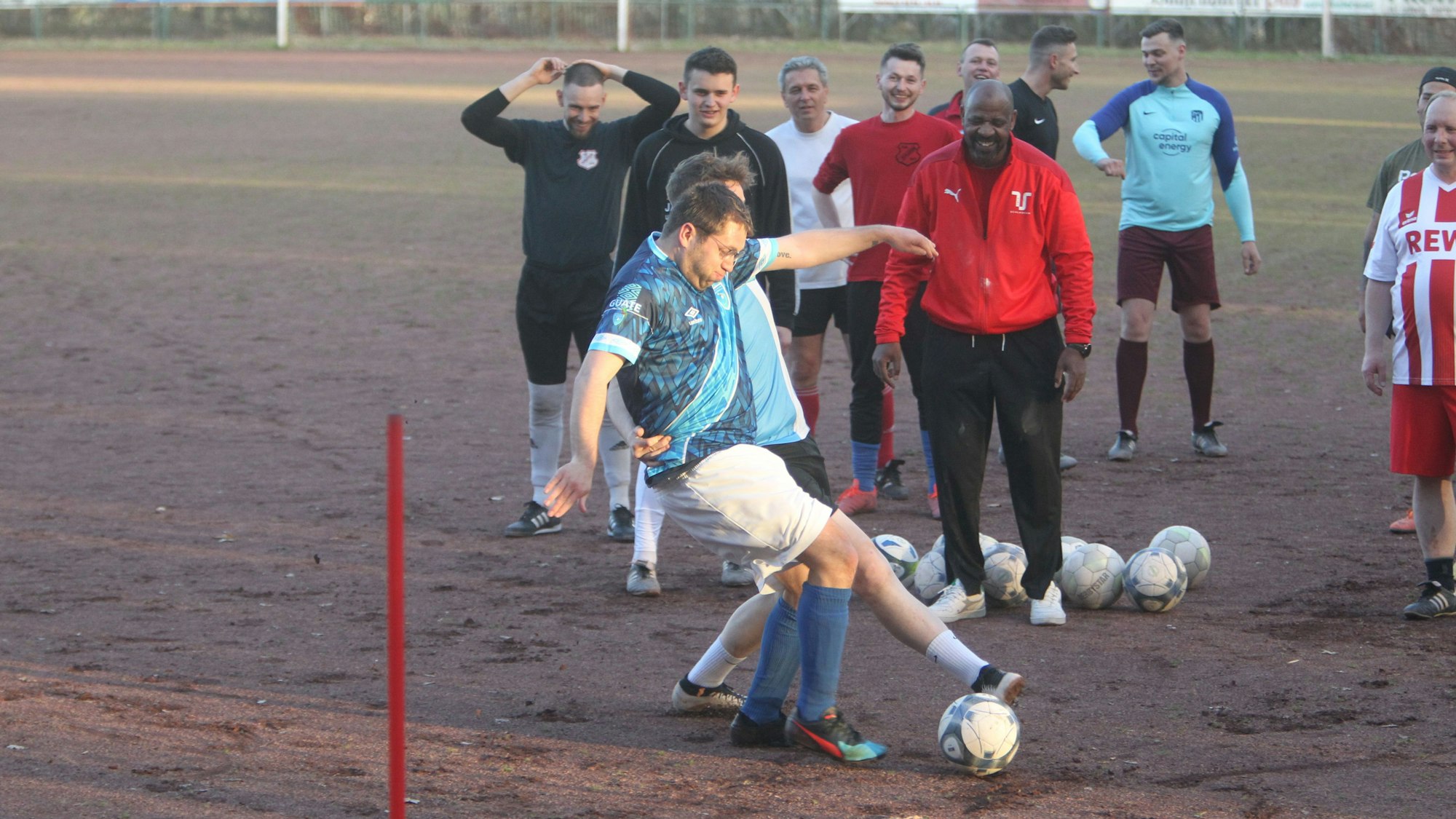 Zwei Fußballer im Ballduell, sie sind von Zuschauern umringt. Der Boden besteht aus roter Asche.