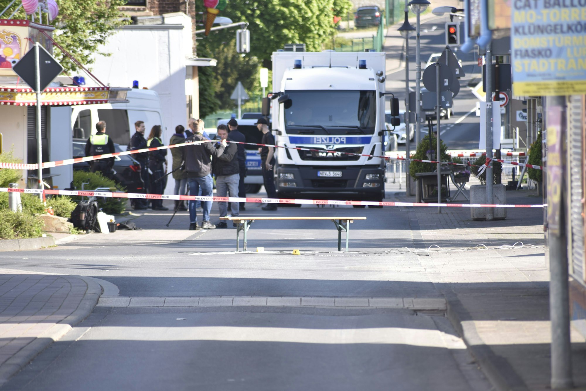 Auf der Kreuzung Weierstraße/Bergstraße in Mechernich steht eine Bierbank, davor sind Markierungen der Polizei zu sehen. Der Bereich drumherum ist mit Flatterband abgesperrt, im Hintergrund sind Polizeiwagen und Kirmesbuden zu sehen.