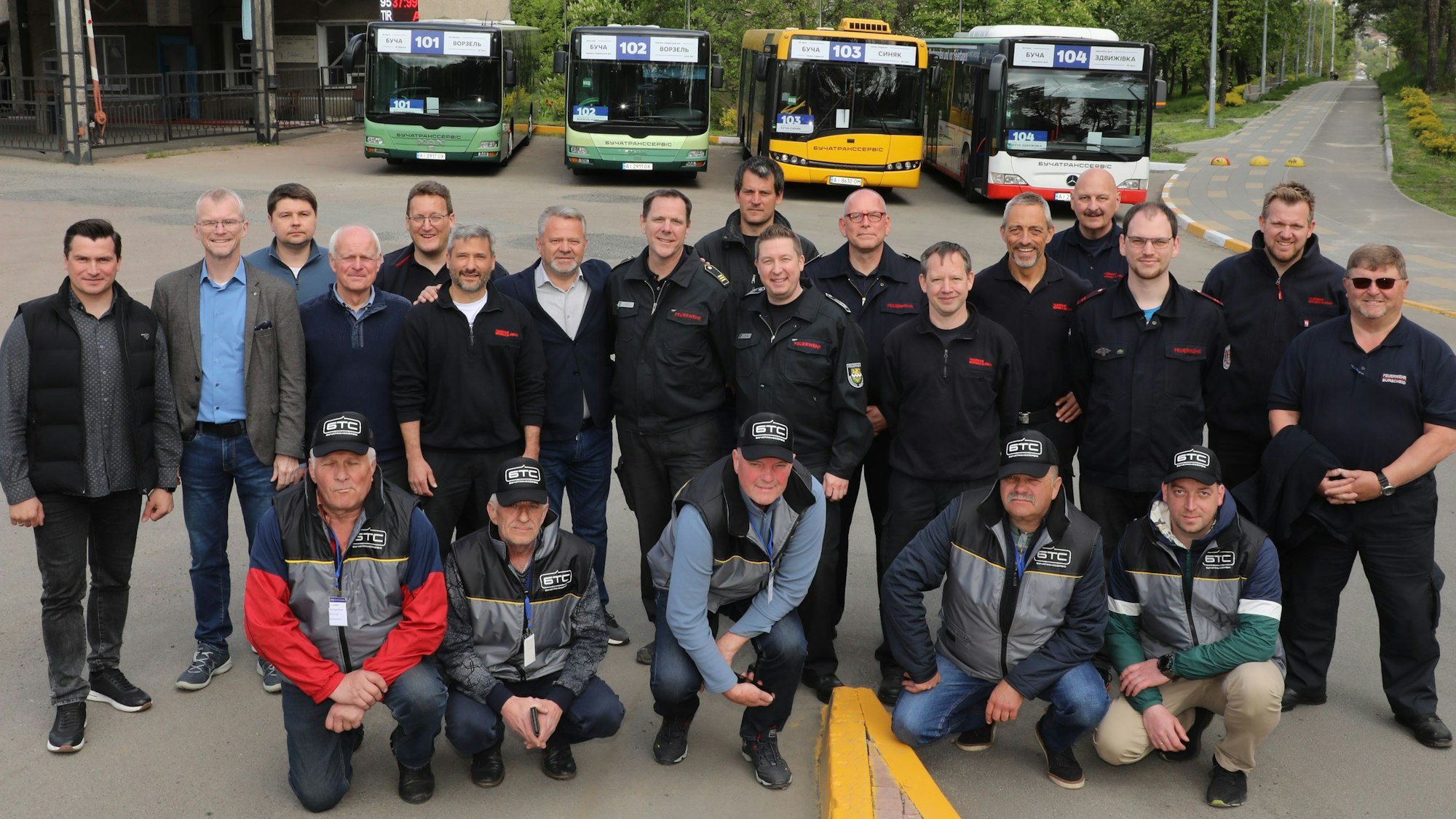 Eine Gruppe von Männern steht beziehungsweise hockt auf einem Platz. Hinter ihnen stehen Busse unterschiedlicher Linien.