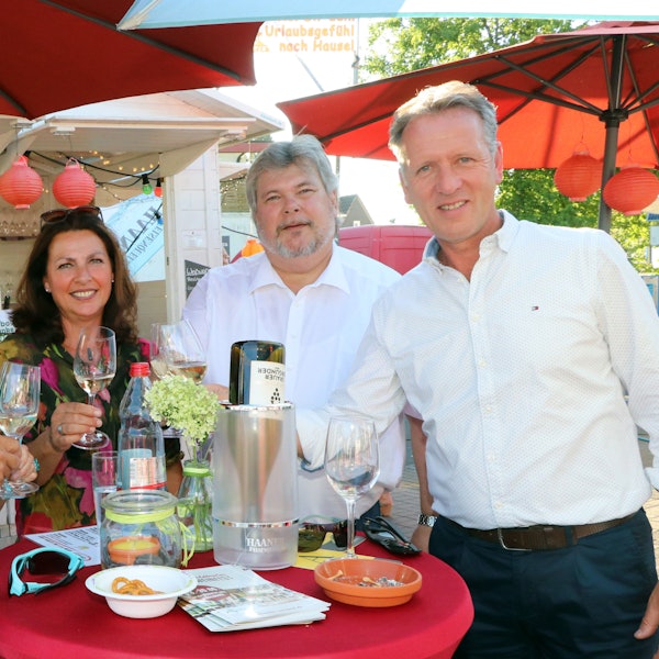 Zwei Männer und eine Frau stehen auf einem Fest an einem Stehtisch und trinken Wein.