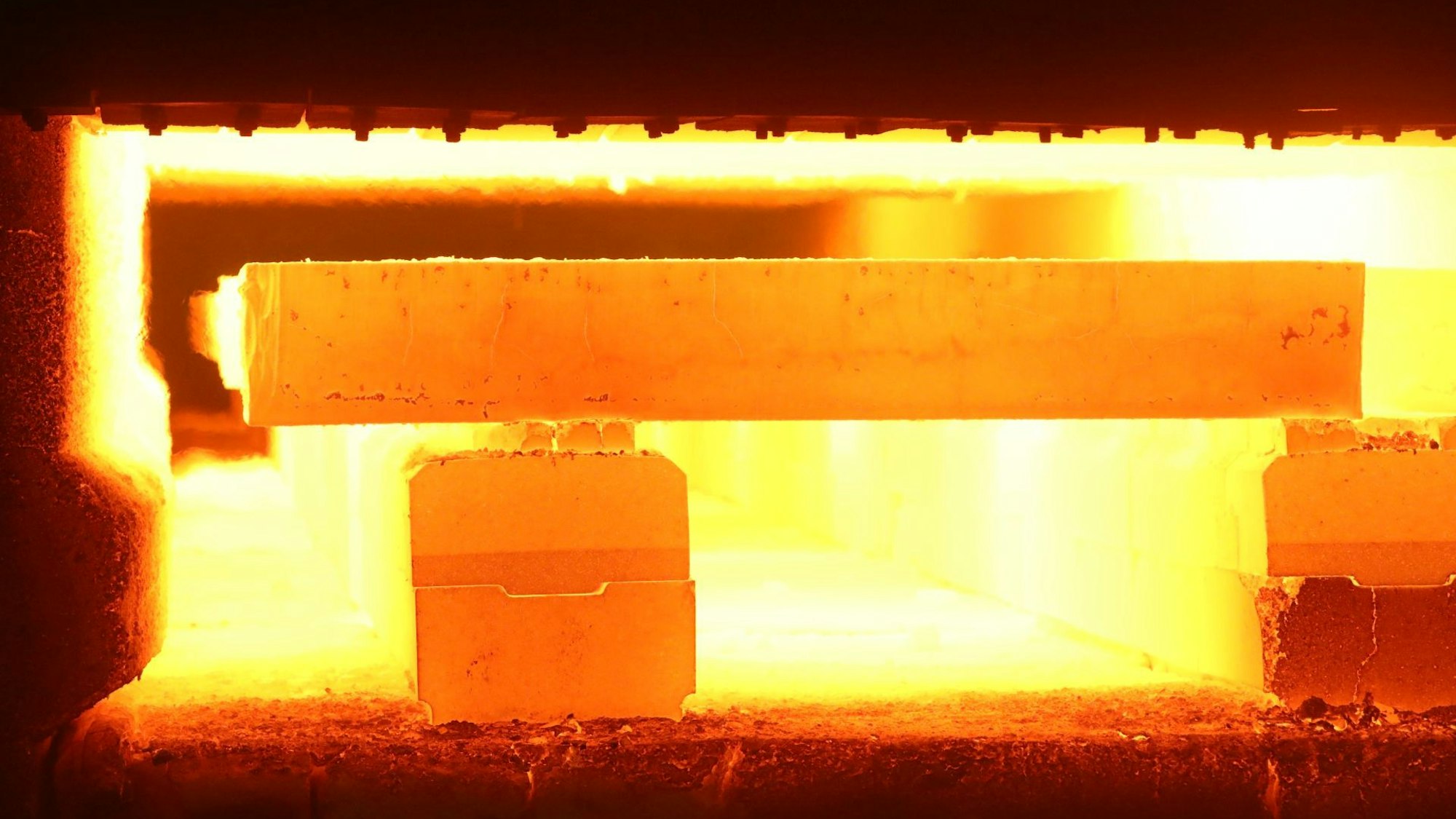 Hinter der kurz geöffneten Klappe eines riesigen Ofens sind rot glühende Stahlblöcke und ein gelber Feuerschein zu sehen.