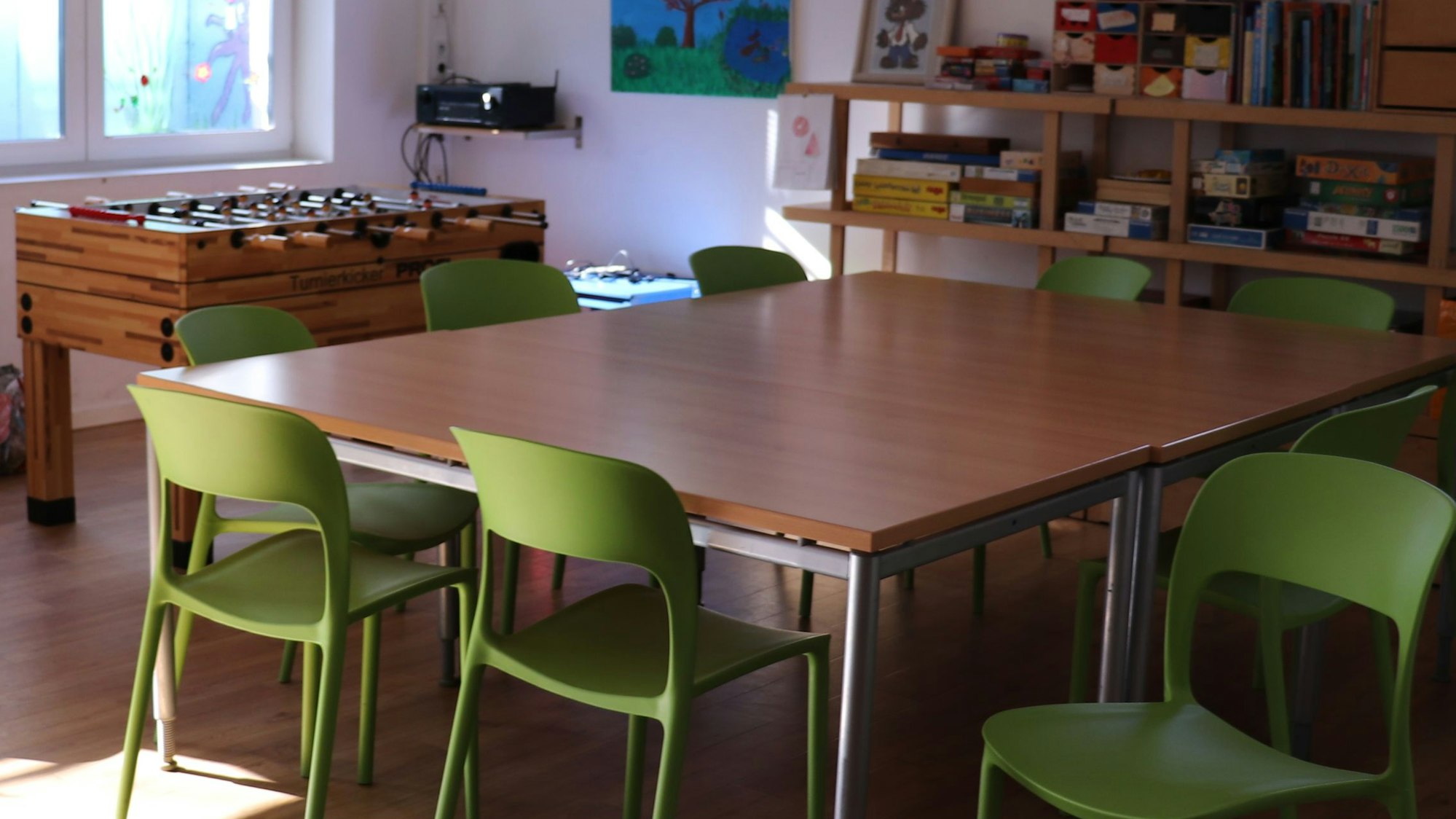 In einem Unterrichtsraum steht ein großer Tisch mit grünen Stühlen, ein Tischkicker und viele Spiele liegen in einem Regal.