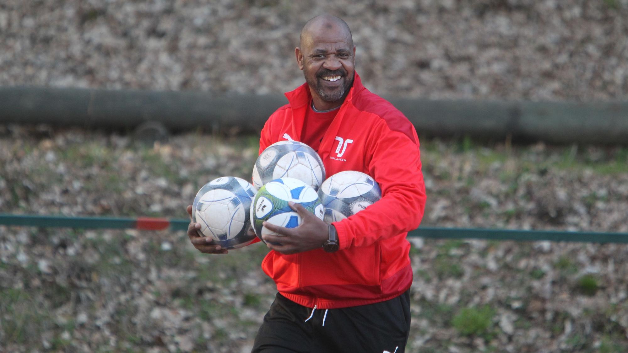 Ein Mann in Trainingsjacke hält vier Fußbälle in den Armen.