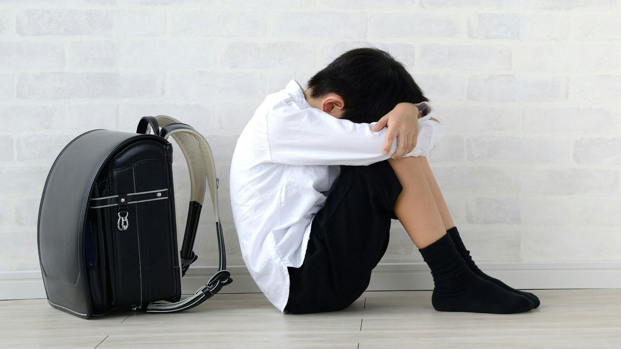 Ein Schuljunge sitzt vor seinem Schulranzen und hält seinen Kopf zwischen den Armen