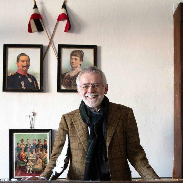 Dr. Peter Joerißen, Leiter Schulmuseum Bergisch Gladbach, im alten Klassenzimmer der ehemaligen Volksschule. Er steht vor einigen Gemälden, die an einer weißen Wand hängen, und lächelt an der Kamera vorbei.
