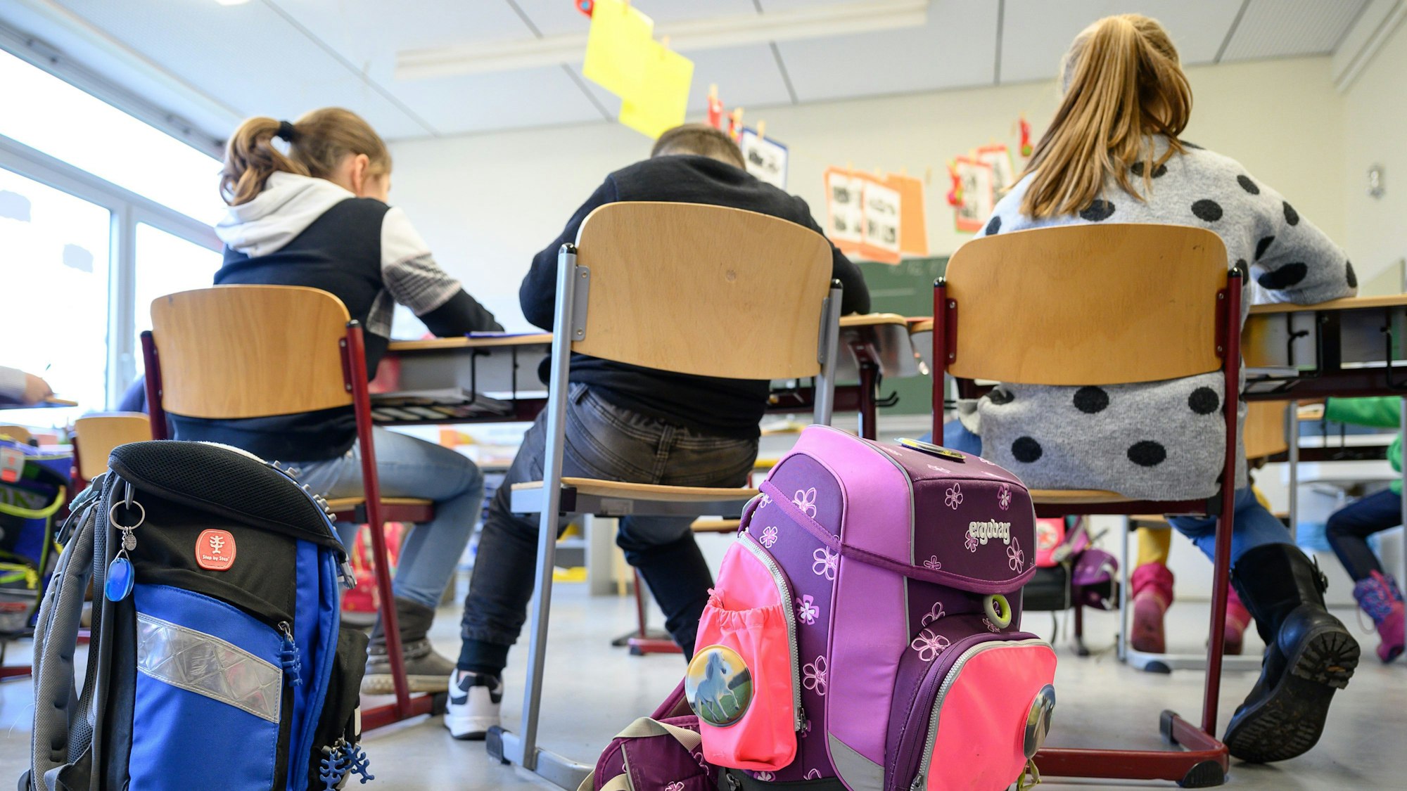 Kinder sitzen auf Stühlen in einem Klassenzimmer. Im Vordergrund sind Schulranzen zu sehen.