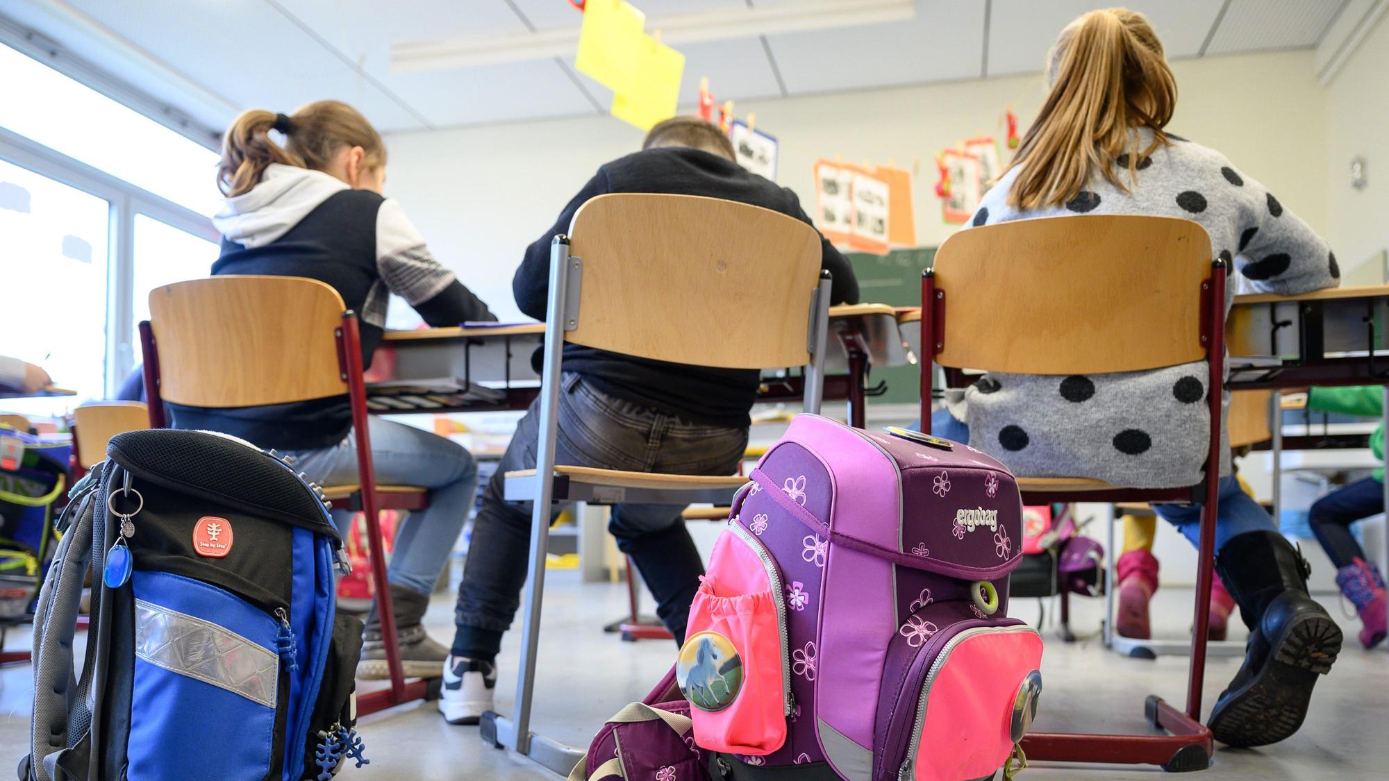 Kinder sitzen in einer Grundschulklasse. Im Vordergrund stehen ihre Ranzen.