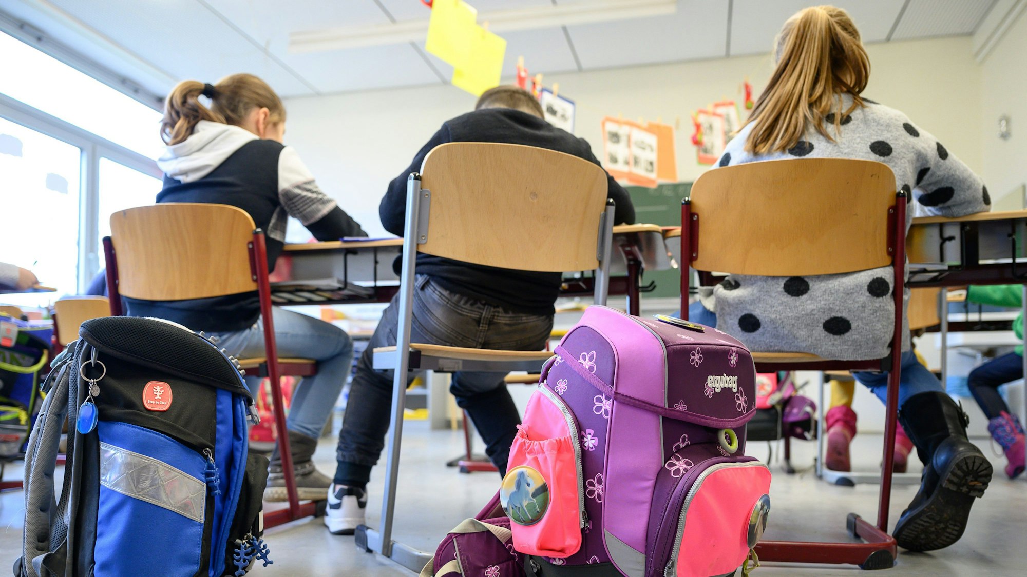 Kinder sitzen auf Stühlen in einem Klassenraum. Im Vordergrund stehen Schulranzen.