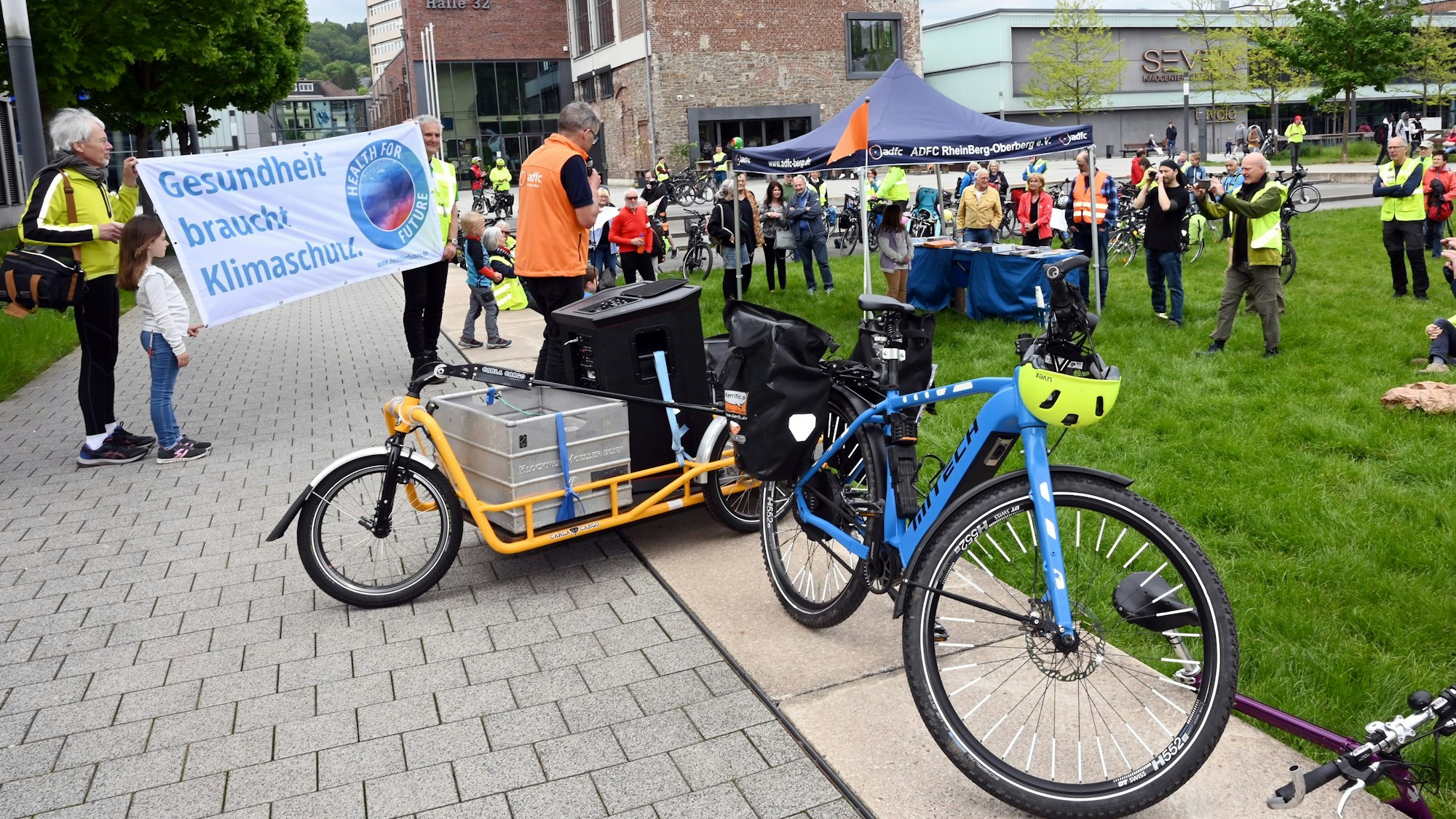 Zu sehen sind in gelbe Westen gekleidete Radfahrer, die sich versammeln und Transparente hochhalten.