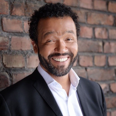 Kabarettist Marius Jung trägt ein weißes Hemd und eine schwarze Anzugjacke und lehnt lächelnd an einer Mauer