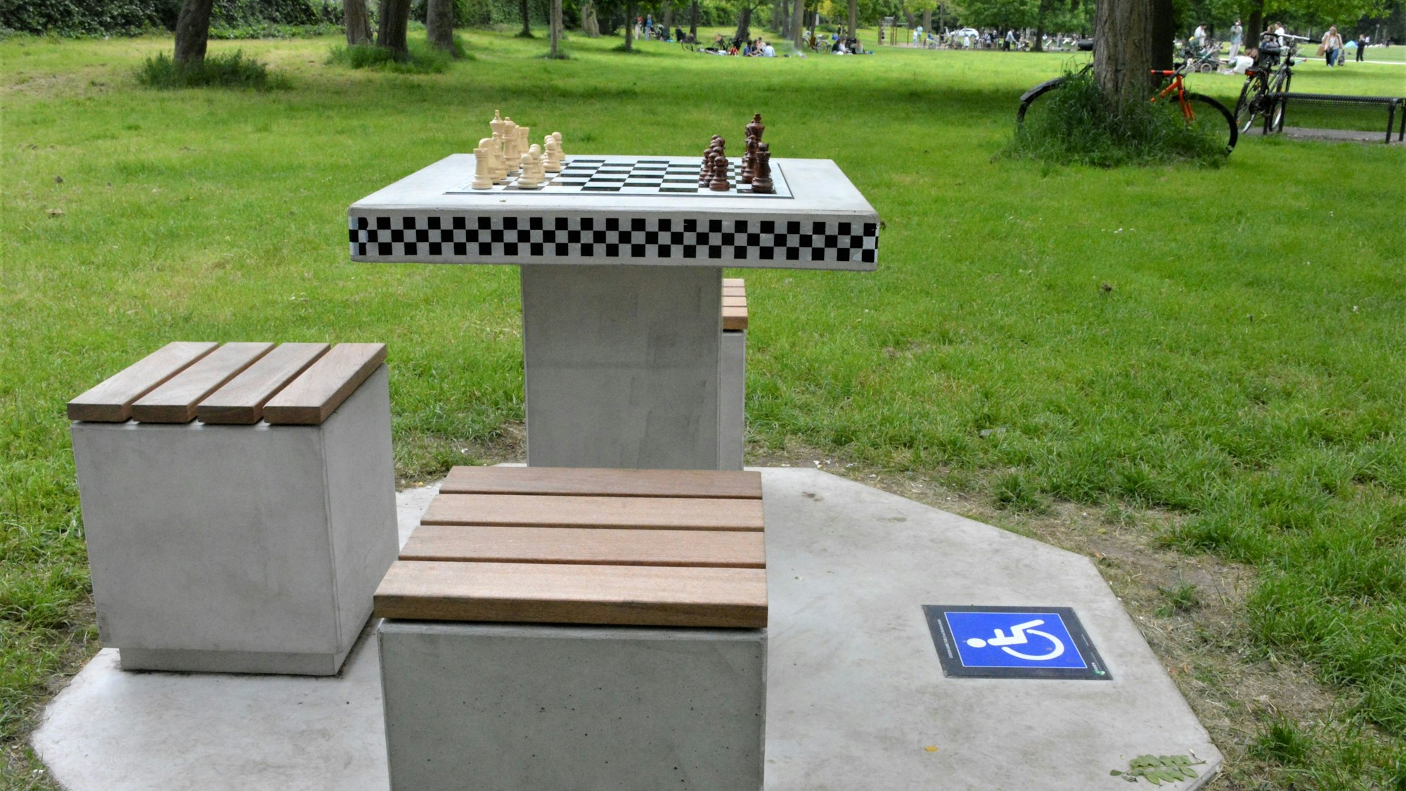 Ein Schachtisch aus Beton steht im Grüngürtel, eine Aufstellfläche für einen Rollstuhl wird mit einem Signet angezeigt.