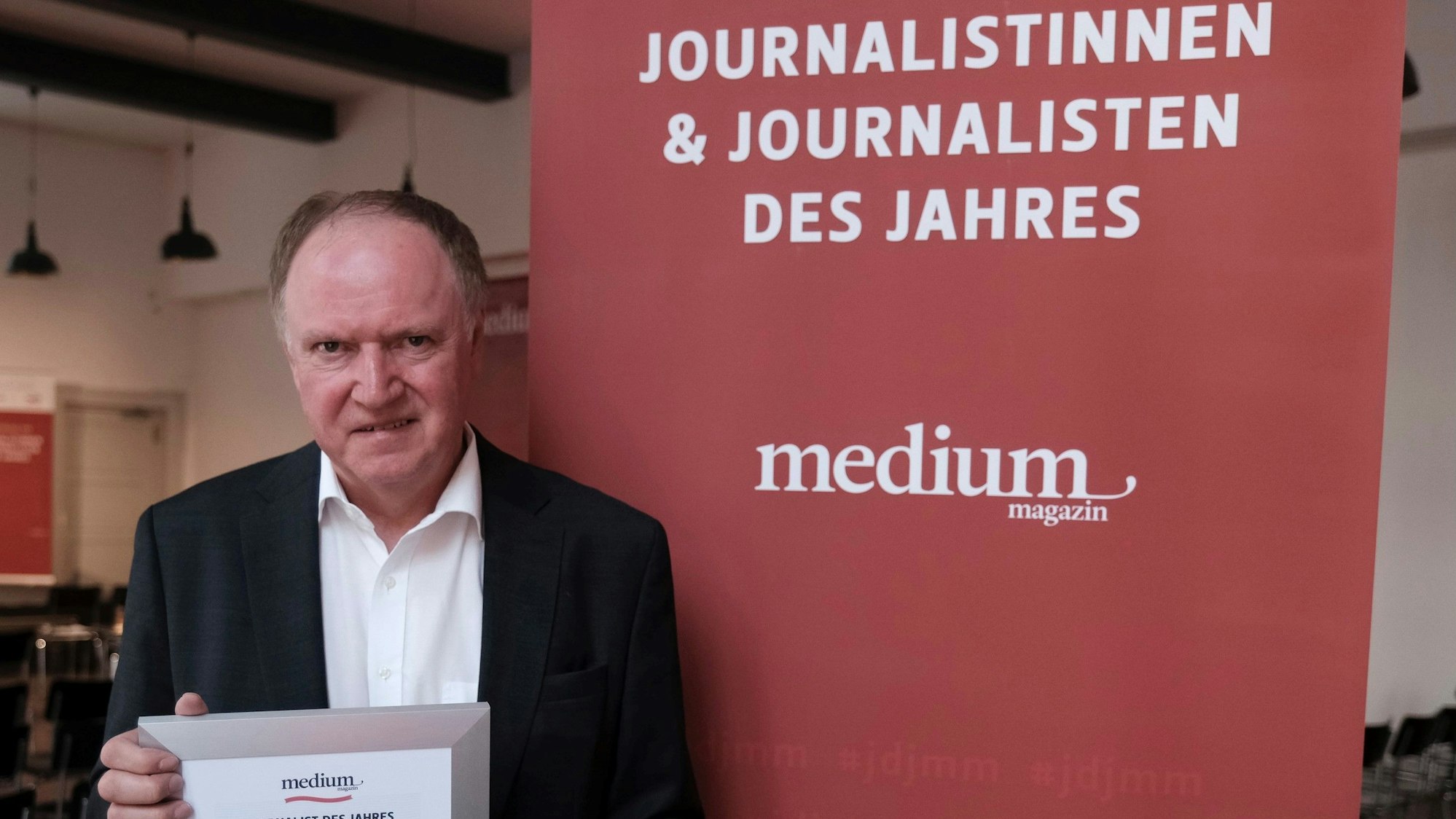 Gerhard Voogt mit seinem Ehrungsdokument steht vor einer roten Info-Wand des Medium Magazins.