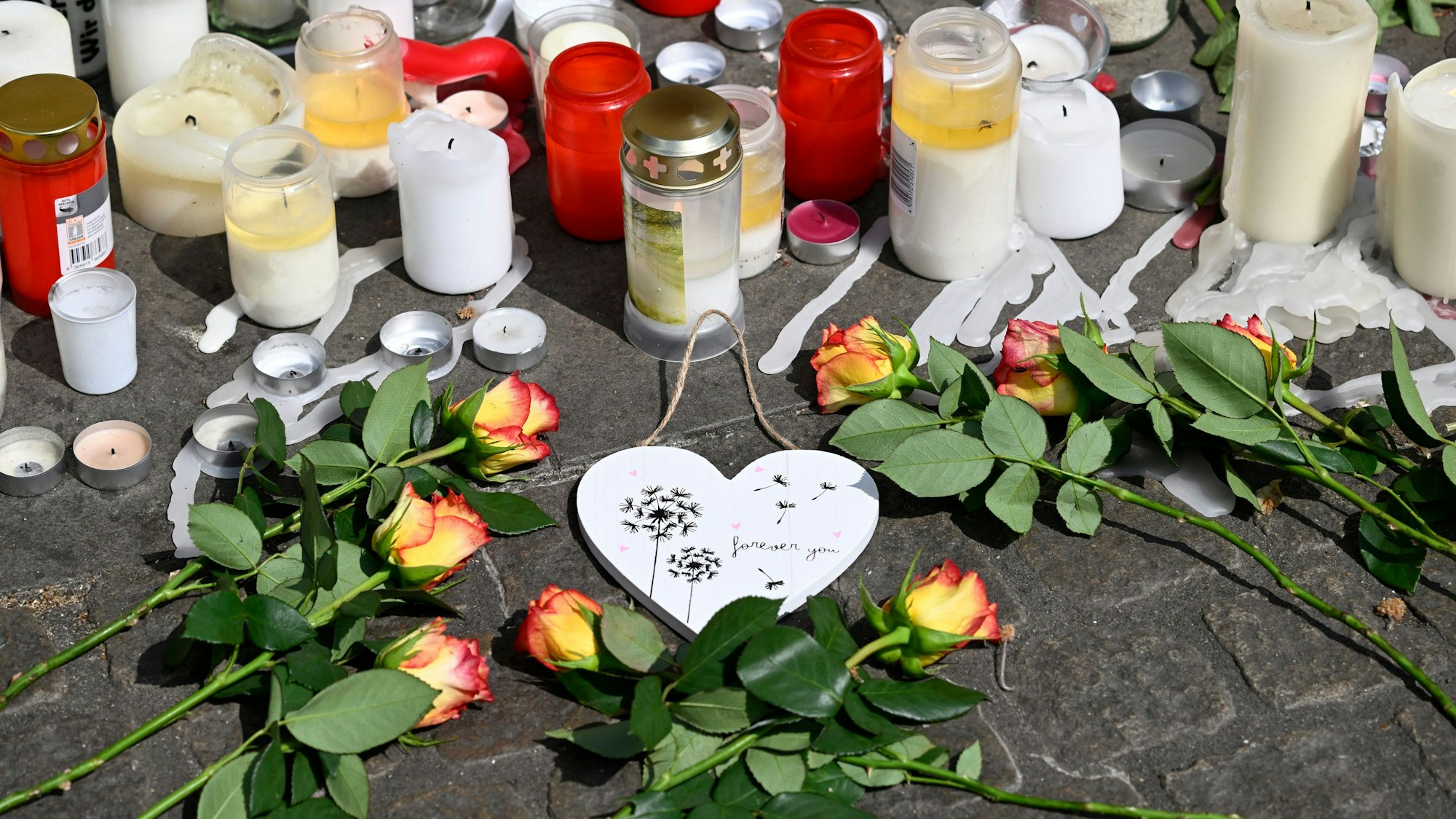 Vor weißen und roten Kerzen liegen ein Holzherz und Blumen.