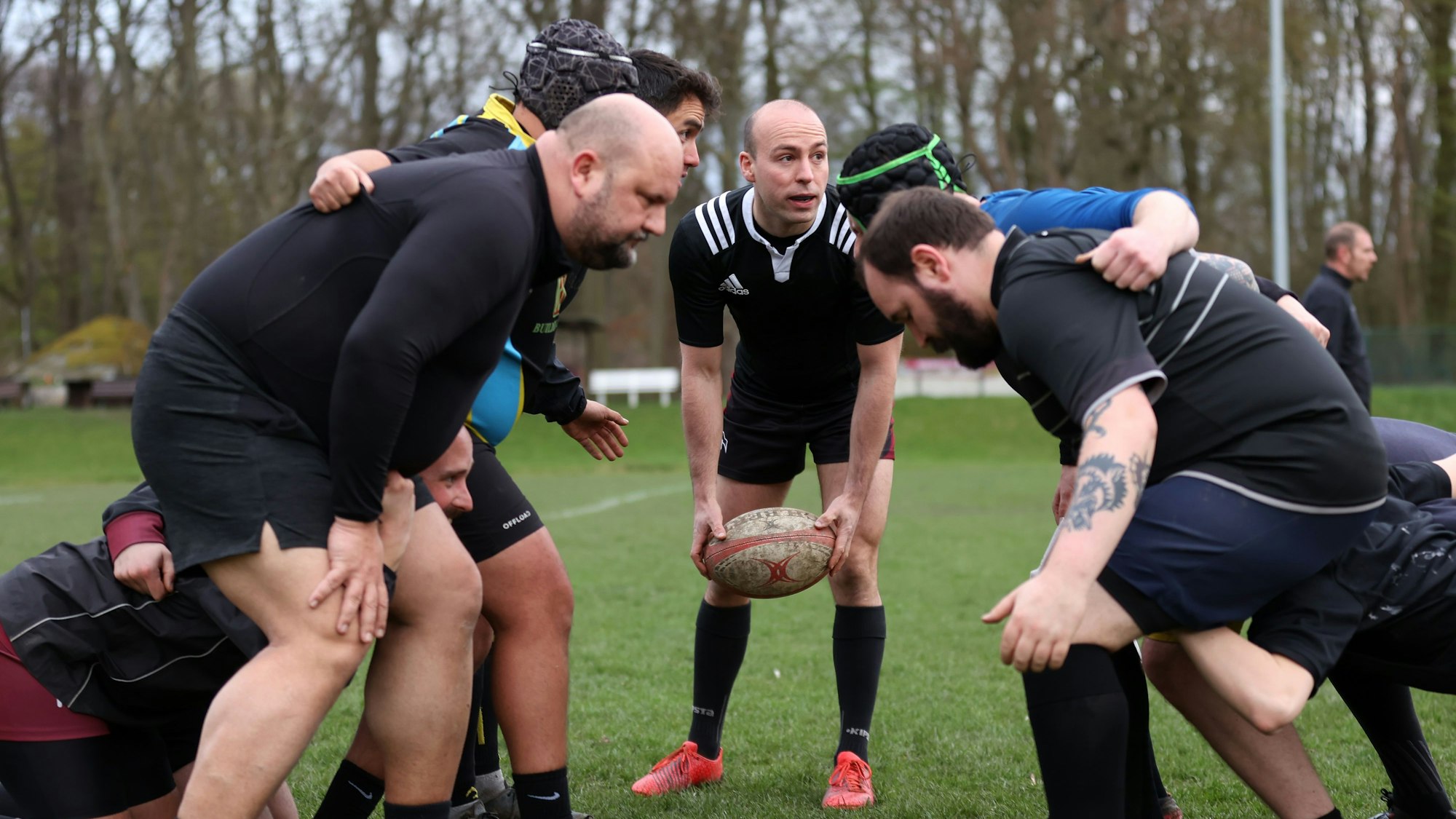 Die queere Rugby-Mannschaft beim Training. Zwei Reihen Männer stehen sich in Startposition gegenüber, der Trainer hält noch den ovalen Ball.