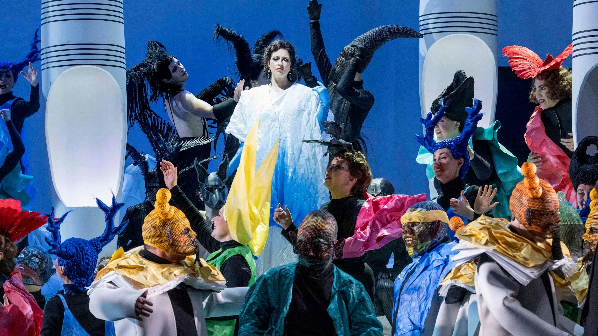 Die Tänzer tragen bunte Kostüme. Im Hintergrund wird eine apathisch blickende Frau in weißen Kleidern von schwarzgekleideten, kuriosen Figuren umzingelt.