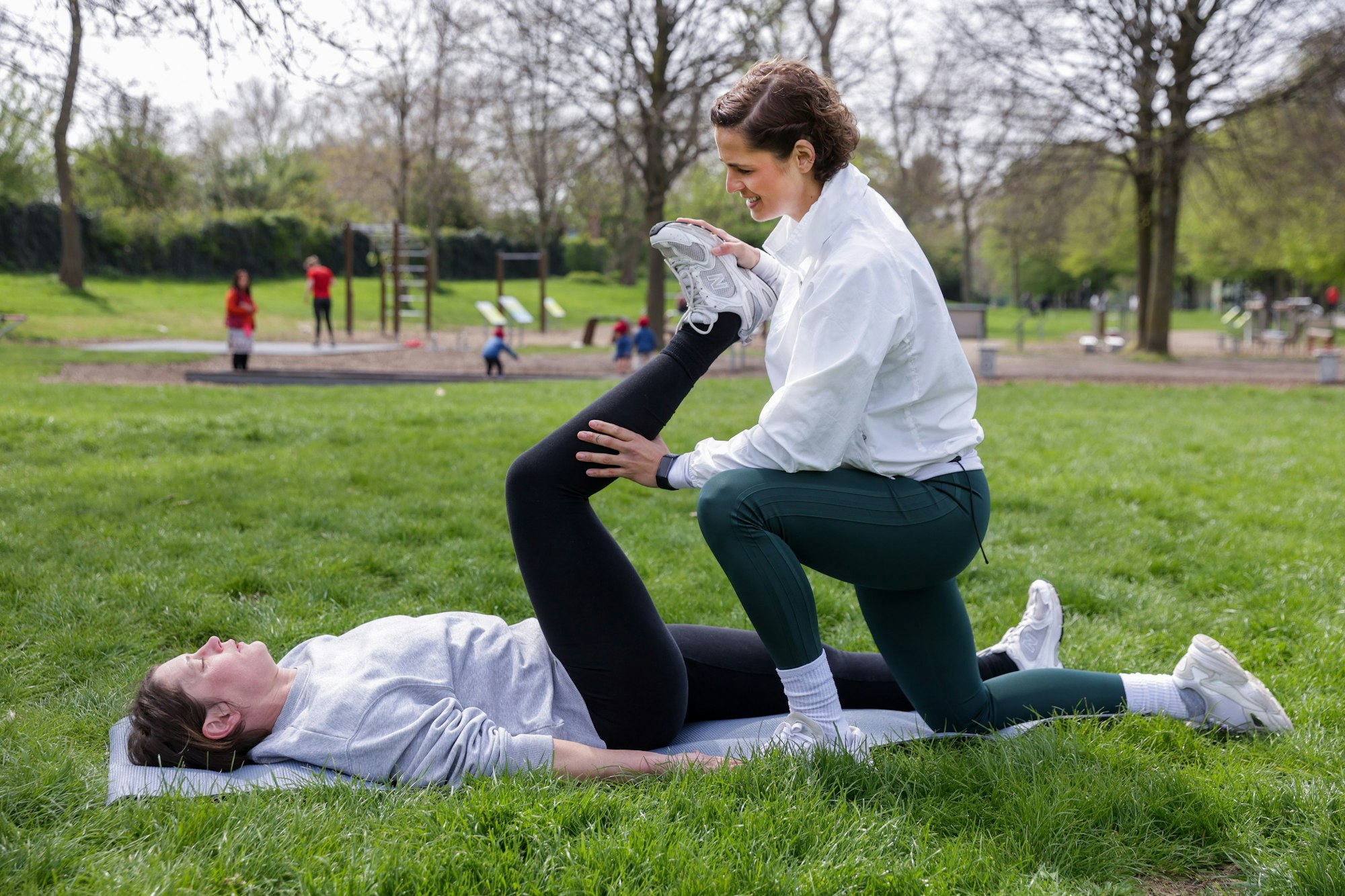 Lara Lueg dehnt das Bein von Paula Engel, die auf dem Rasen im Park liegt.