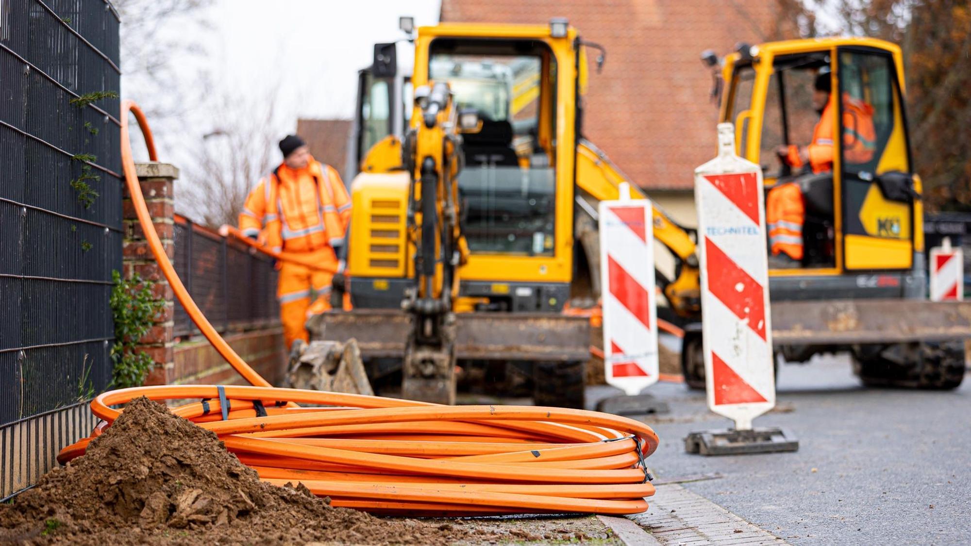 Arbeiter sind mit Bagger und Spaten an einer Straße mit der Erdverlegung von Glasfaserkabeln beschäftigt.