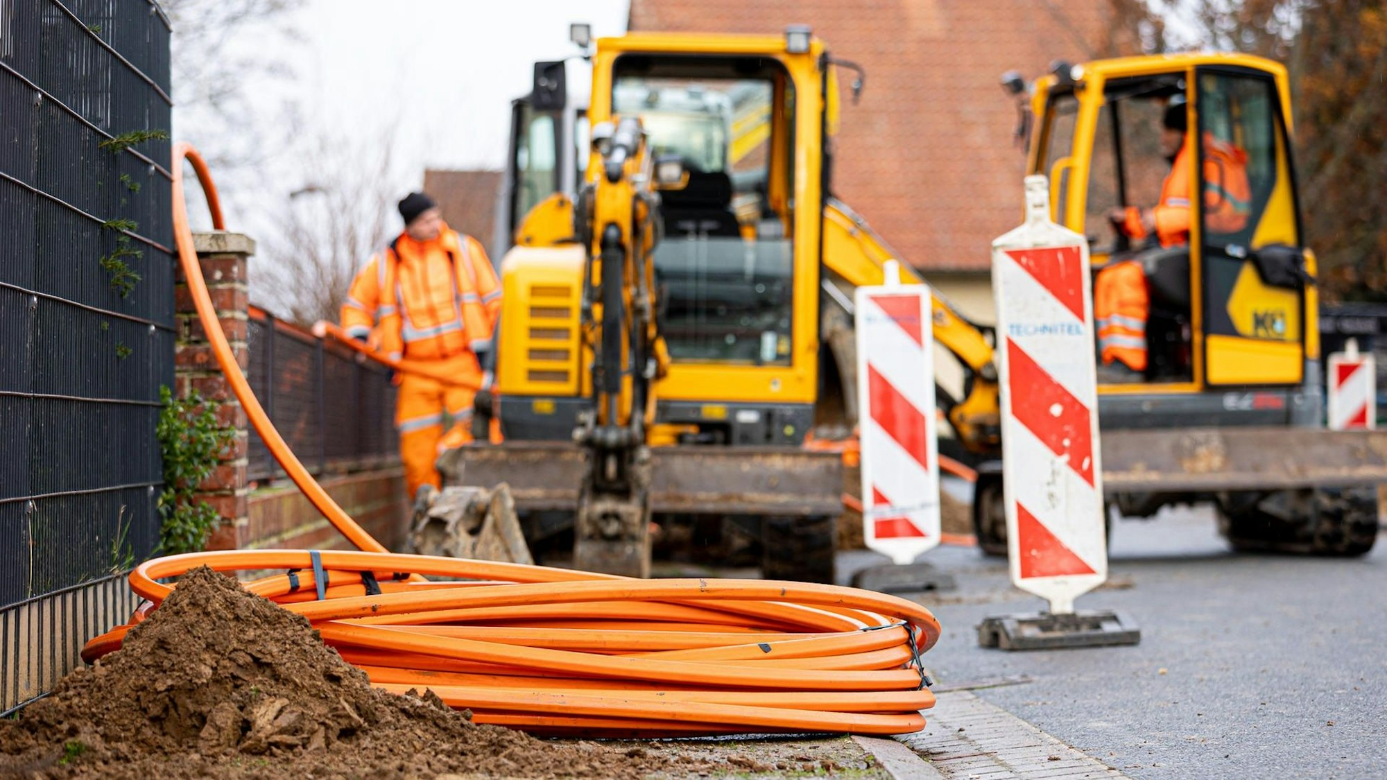 Arbeiter sind mit Bagger und Spaten an einer Straße mit der Erdverlegung von Glasfaserkabeln beschäftigt. (Symbolbild)