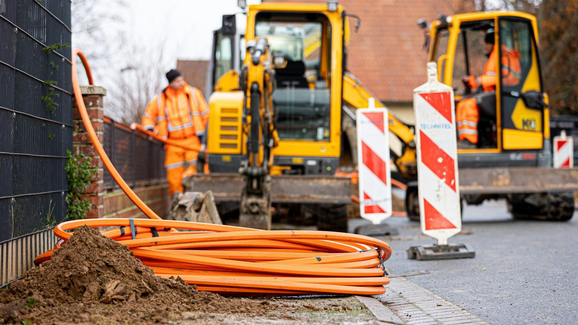 Arbeiter sind mit Bagger und Spaten an einer Straße mit der Erdverlegung von Glasfaserkabeln beschäftigt.