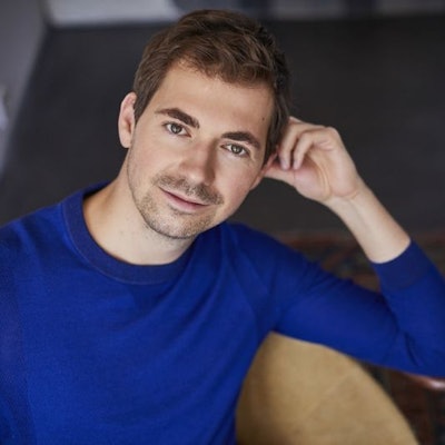 Valer Sabadus trägt einen blauen Pullover. Er hat braune Haare und einen 3-Tage-Bart. Er sitzt und stützt sich mit dem Ellbogen an der Stuhllehne ab.