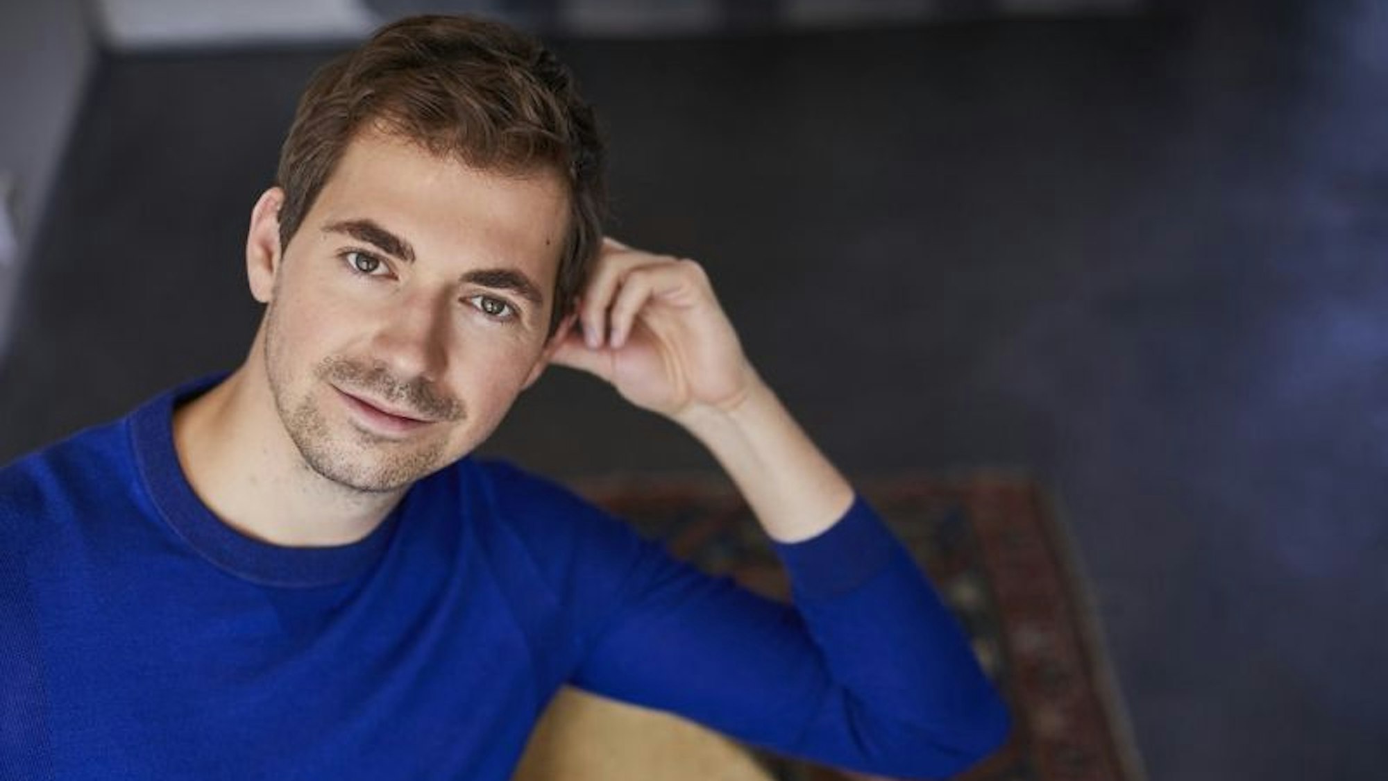 Valer Sabadus trägt einen blauen Pullover. Er hat braune Haare und einen 3-Tage-Bart. Er sitzt und stützt sich mit dem Ellbogen an der Stuhllehne ab.