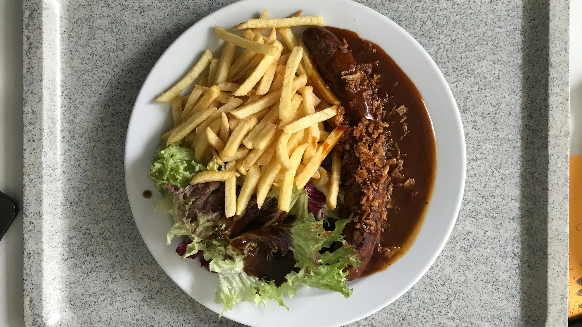Ein Teller mit Currywurst, Pommes frites und einer kleinen Salatbeilage steht auf einem Tablet in einer Kantine.