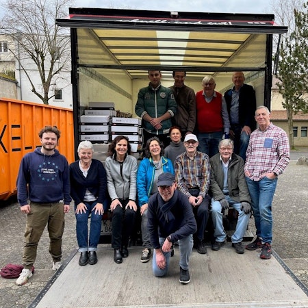 Eine Gruppe von Männern und Frauen stehen beziehungsweise sitzen am geöffneten Laderaum eines Lastwagens.