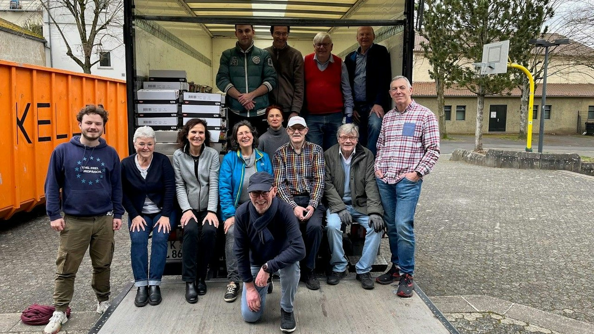 Eine Gruppe von Männern und Frauen stehen beziehungsweise sitzen am geöffneten Laderaum eines Lastwagens.
