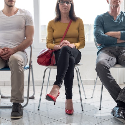 Drei Menschen sitzen nebeneinander in einem Raum auf Stühlen und warten.