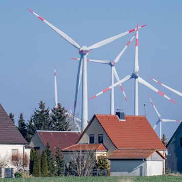ARCHIV - 01.03.2023, Sachsen-Anhalt, Wethau: Windräder drehen sich hinter einer Siedlung mit Eigenheimen bei Wethau in Sachsen-Anhalt. Strom und Gas werden für einige Haushaltskunden etwas günstiger - allerdings oft auf hohem Niveau.