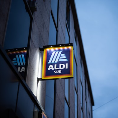 Das Logo einer Aldi Süd Filiale, fotografiert kurz vor dem Sonnenuntergang. Das Schild wird beleuchtet.