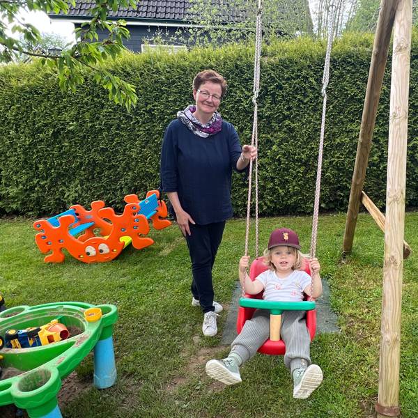Eine Frau mit Brille steht hinter einem schaukelnden Kind mit Kappe.