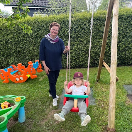 Eine Frau mit Brille steht hinter einem schaukelnden Kind mit Kappe.