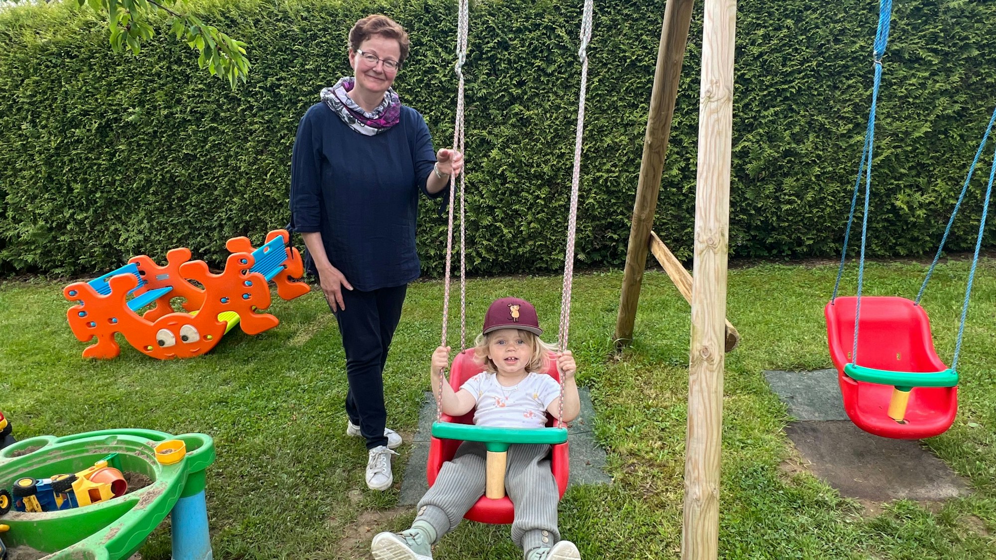 Es ist Martha Göttfert mit ihrem Tageskind Neni zu sehen. Das Kind sitzt auf einer Schaukel, Martha steht daneben.