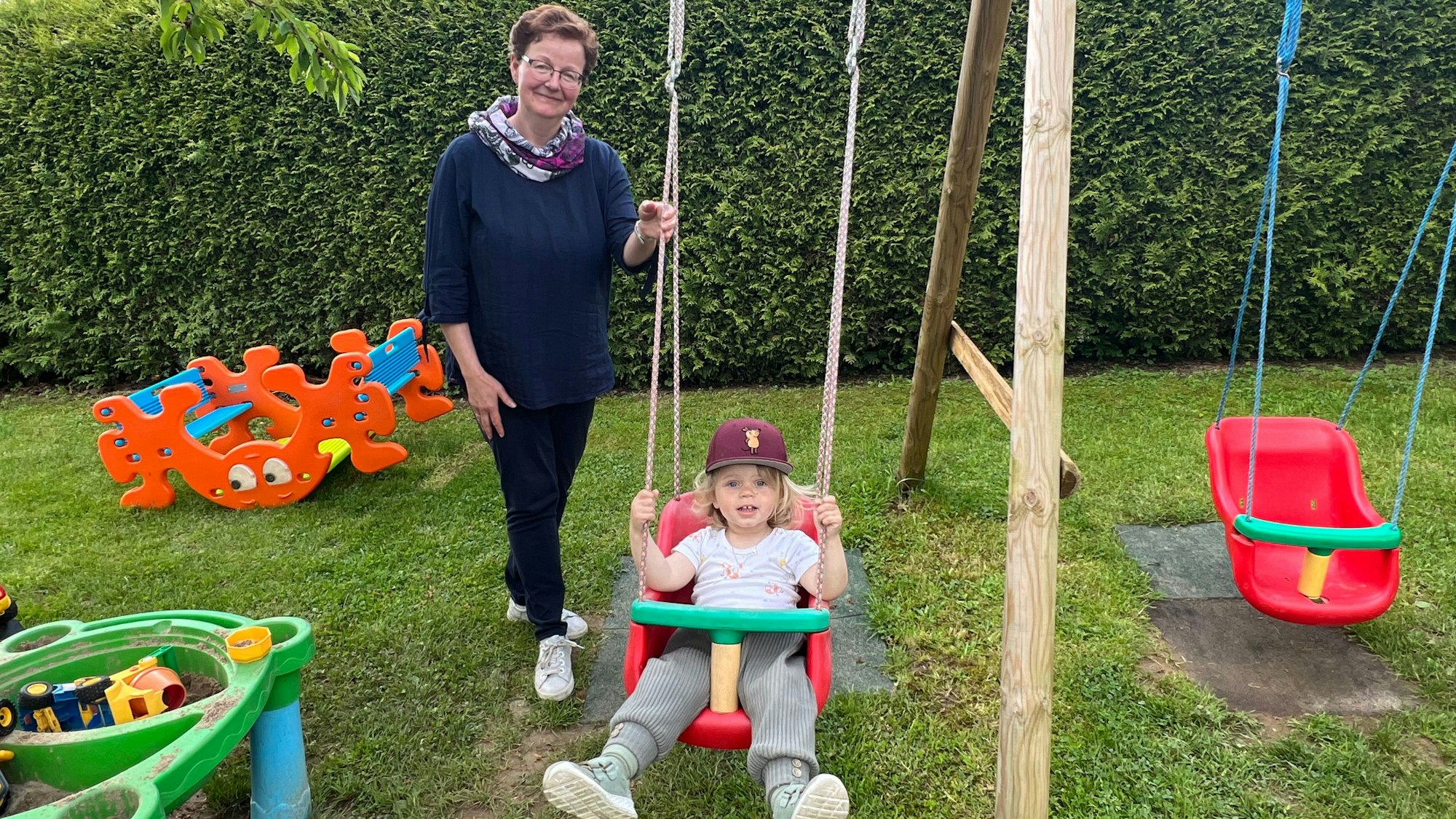 Eine Frau mit Brille steht hinter einem schaukelnden Kind mit Kappe.