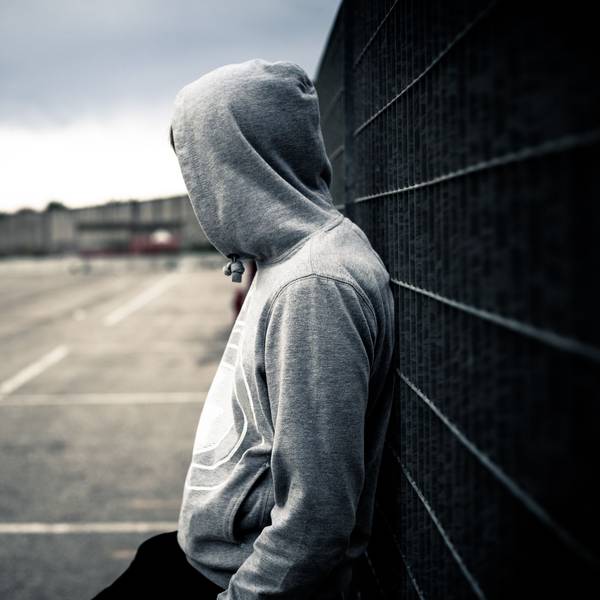 Ein junger Mann mit Kapuzenpulli lehnt alleine an einem Zaun.