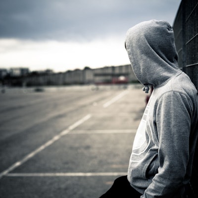 Ein junger Mann mit Kapuzenpulli lehnt alleine an einem Zaun.