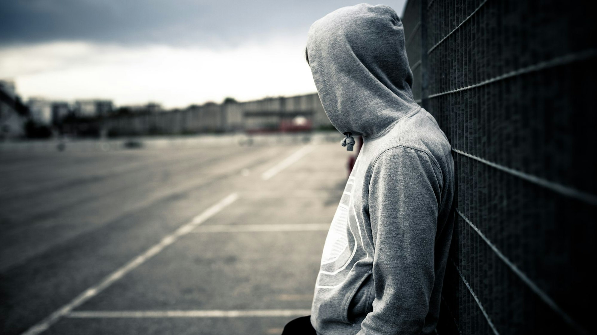 Ein junger Mann mit Kapuzenpulli lehnt alleine an einem Zaun.