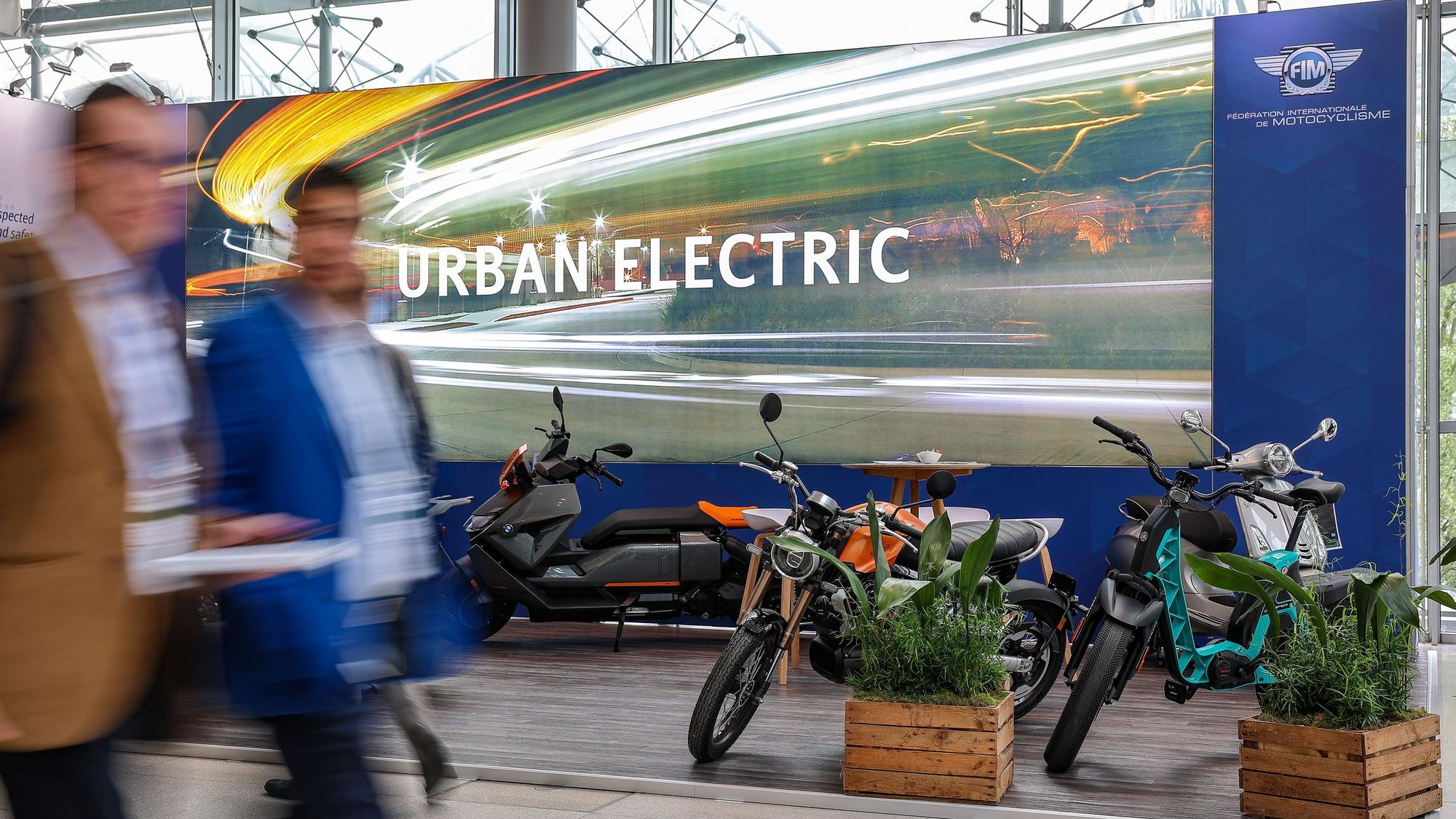 Teilnehmer des Weltgipfels der Verkehrsminister gehen vor einem Stand mit elektrisch angetriebenen Motorrollern und Motorrädern entlang.