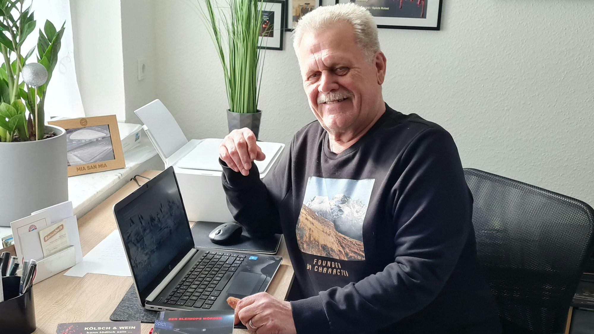 Ein älterer Mann mit Schnurrbart sitzt im Pullover vor einem Laptop und grinst in die Kamera.