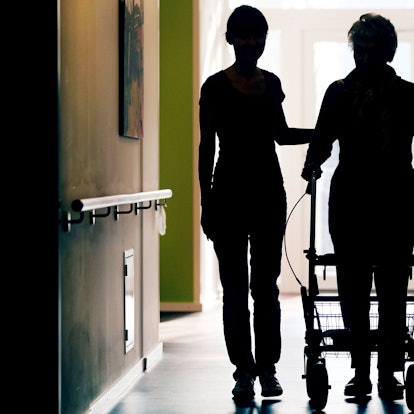 Eine Pflegekraft (l) begleitet die Bewohnerin eines Altenheims beim Gang über den Flur.