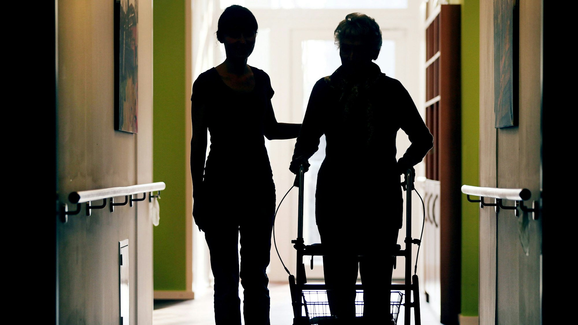 Eine Pflegekraft (l) begleitet die Bewohnerin eines Altenheims beim Gang über den Flur.