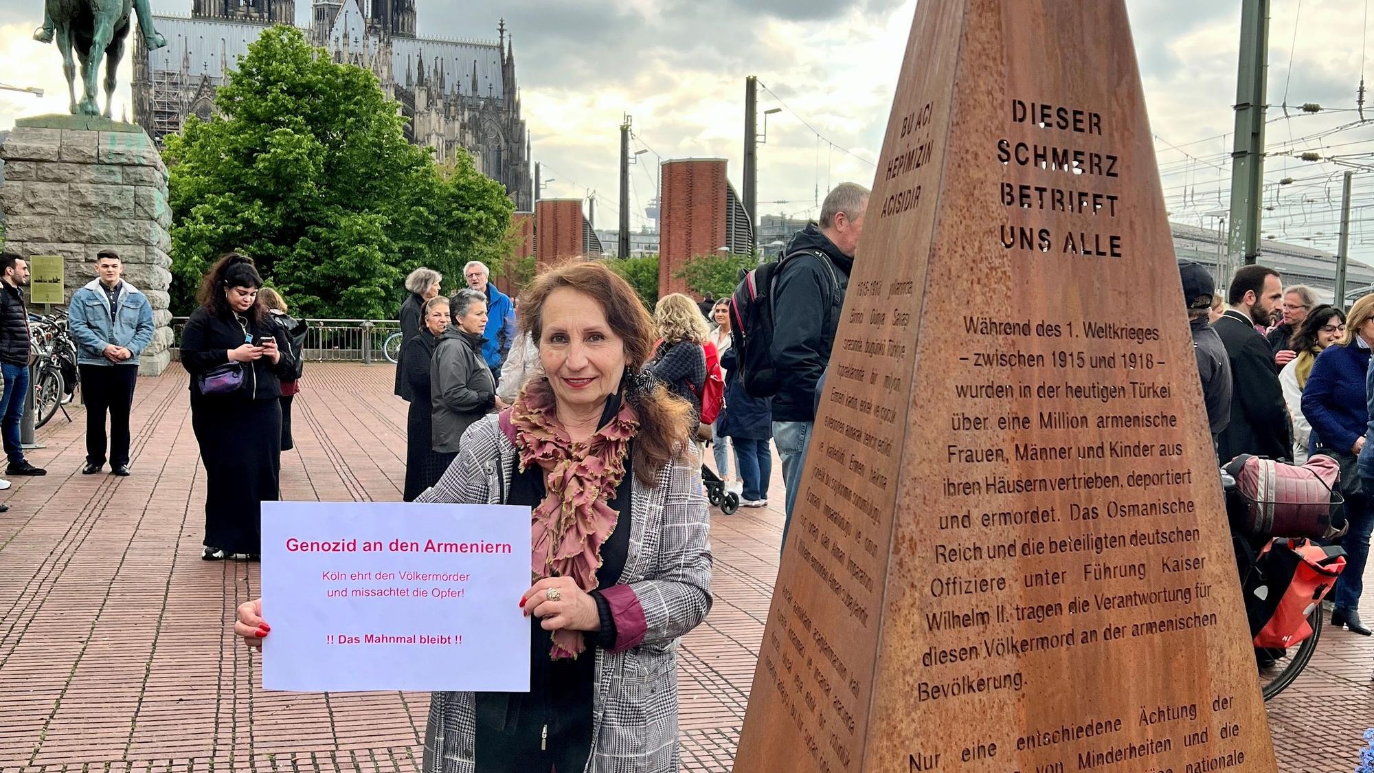 Eine Demonstrantin hält bei einer Kundgebung vor dem Mahnmals für den Völkermord in Armenien an der Hohenzollernbrücke ein Protest-Schild in die Kamera.