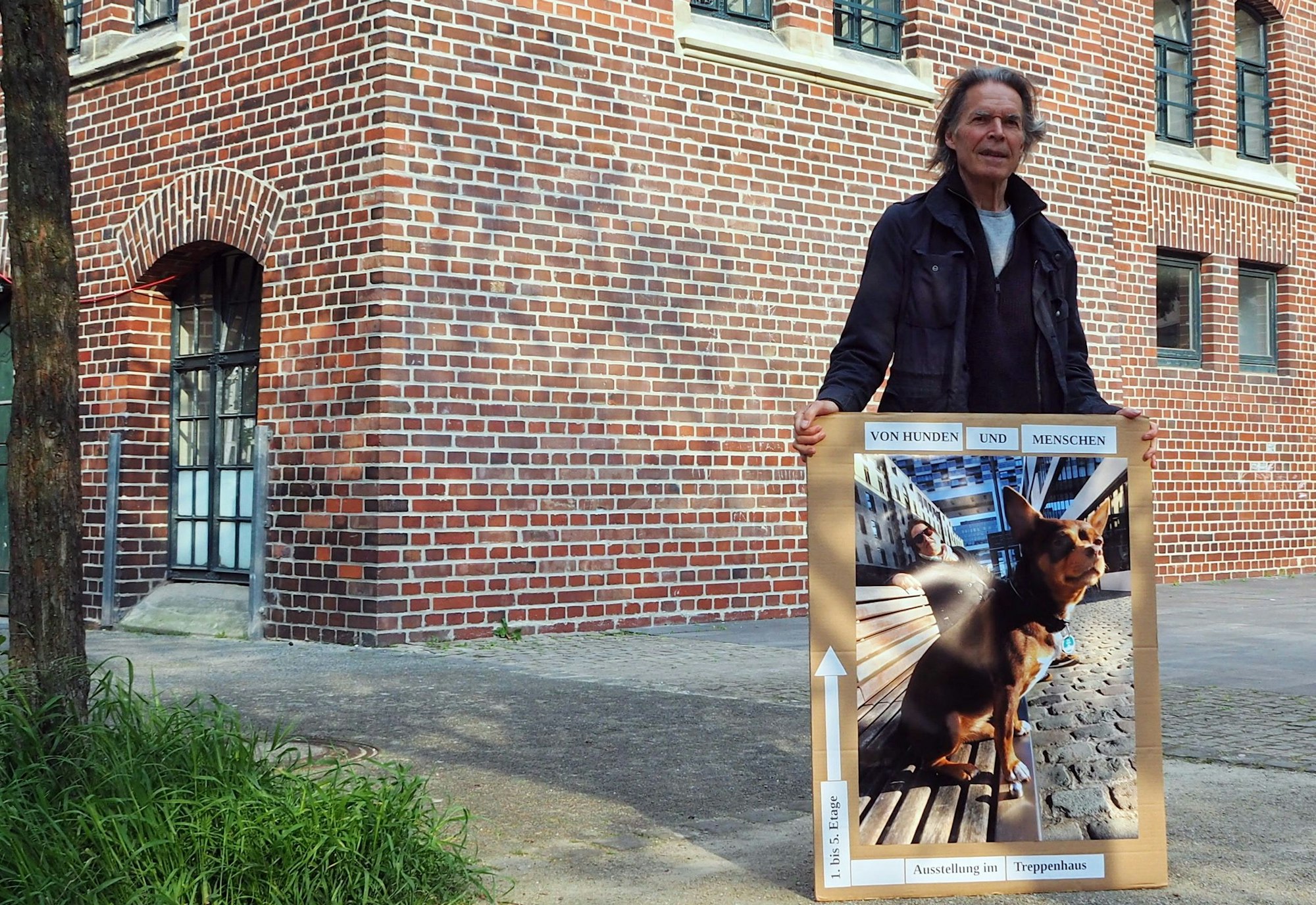 Ein Mann steht mit einem Plakat, das den Weg zur Ausstellung weist, vor einem Backsteinbau, dem Bürgerhaus Stollwerck.